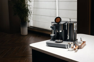 Black Meraki espresso machine with a round touchscreen, beside a stainless steel milk pitcher, wooden-handled portafilter and tamping tools on a white counter.
