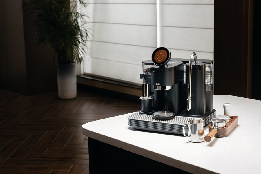 Black Meraki espresso machine with a round touchscreen, beside a stainless steel milk pitcher, wooden-handled portafilter and tamping tools on a white counter.