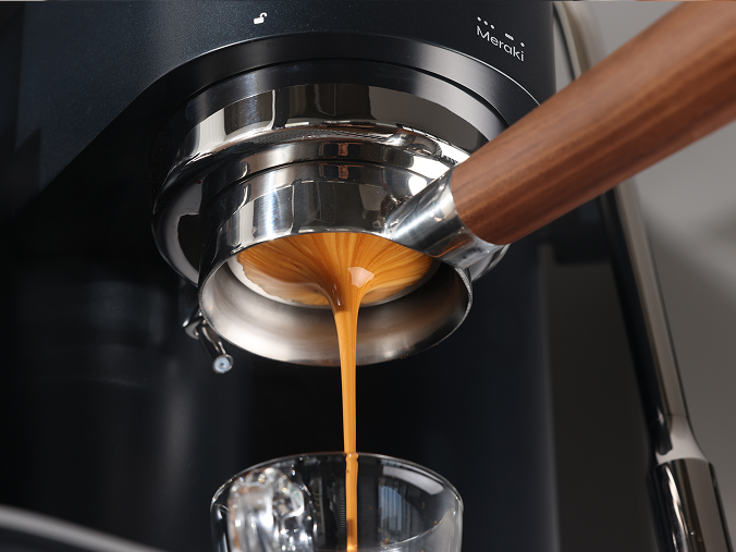 Espresso flowing from a wooden-handled bottomless portafilter into a glass on a black Meraki espresso machine.