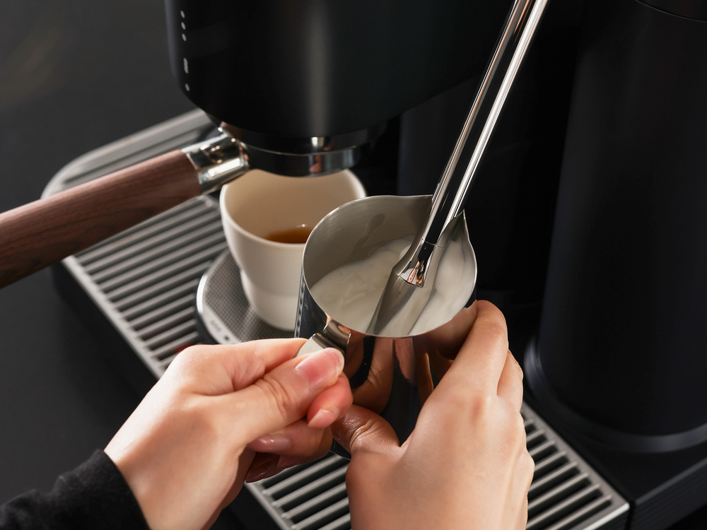 Hands holding a stainless steel pitcher while steaming milk on a Meraki espresso machine, with a cup of espresso beside it.
