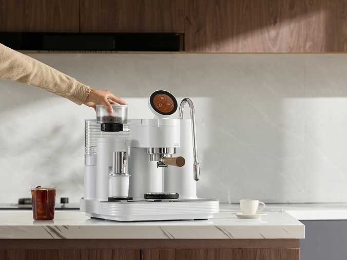 A white coffee machine sitting on a kitchen counter, with a hand adjusting the bean hopper on top, next to an iced Americano and a small espresso cup.